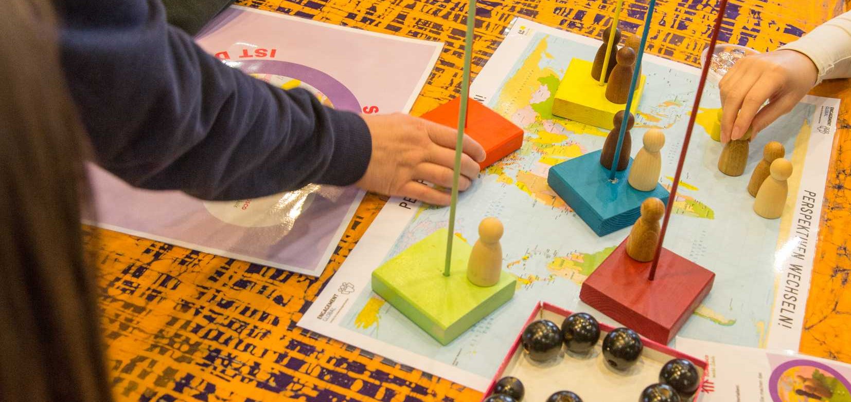 Menschen spielen ein Brettspiel auf einer Weltkarte mit Holzfiguren, farbigen Blöcken und Stäben, um globale Perspektiven zu erkunden.