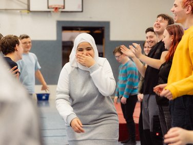 Eine junge Frau mit Hidschab lacht und bedeckt ihren Mund, umgeben von lächelnden und klatschenden Jugendlichen in einer Turnhalle.