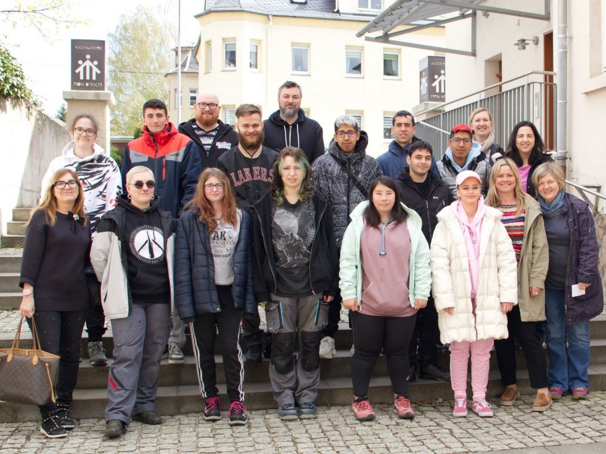 Eine diverse Gruppe von etwa 20 Erwachsenen und Jugendlichen posiert lächelnd auf Außentreppen vor einem Gebäude.