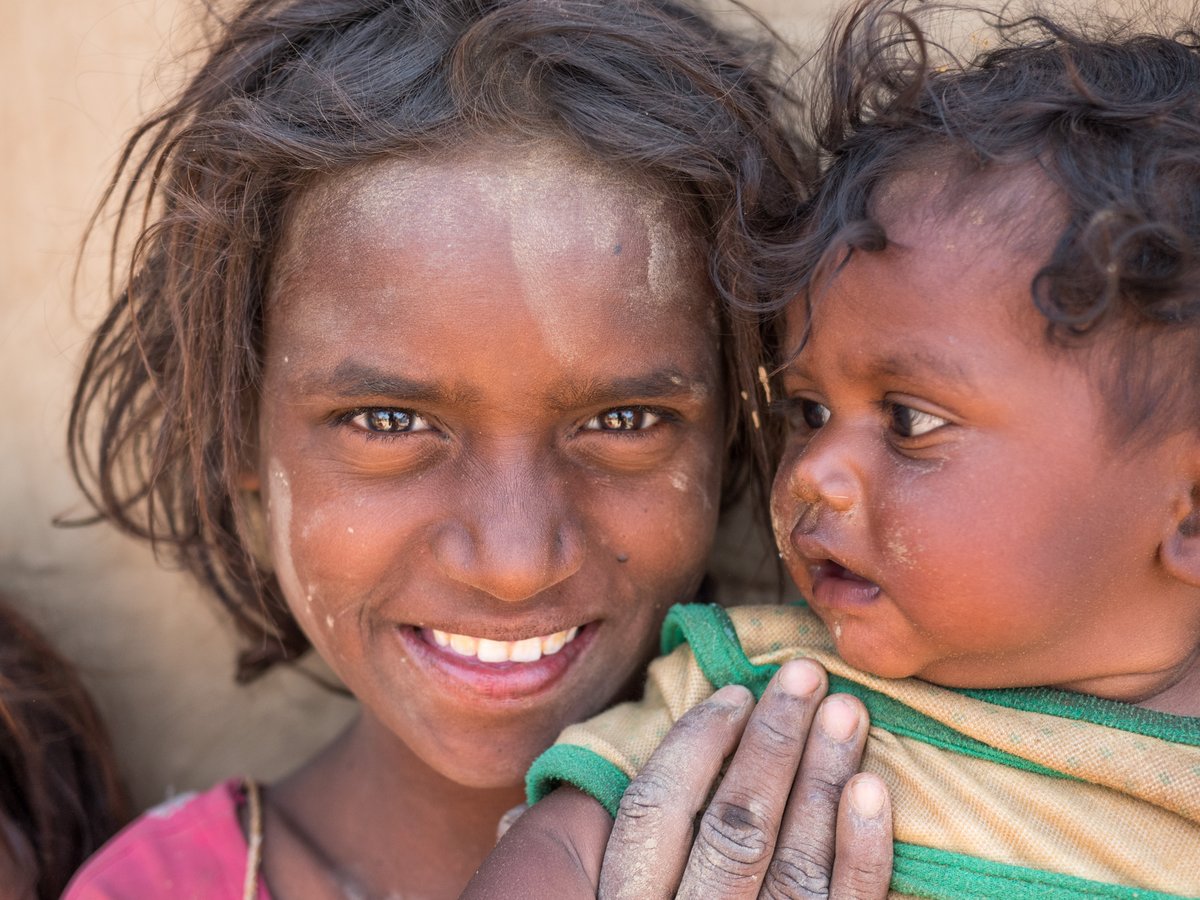 Nahaufnahme von zwei Kindern; ein älteres Mädchen lächelt, hält ein Baby, das nach rechts schaut. Beide staubig.