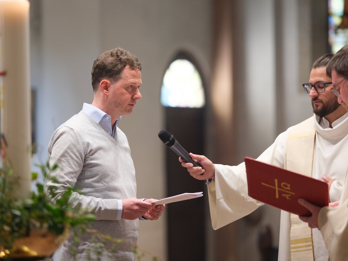 Ein Mann spricht in ein Mikrofon, das von einem Priester gehalten wird, mit einem weiteren Priester daneben, während einer Kirchenzeremonie.