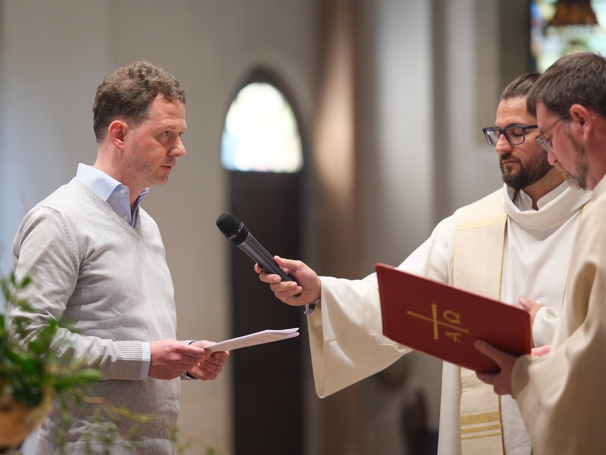 Ein Mann spricht in ein Mikrofon, das von einem Priester gehalten wird, mit einem weiteren Priester daneben, während einer Kirchenzeremonie.