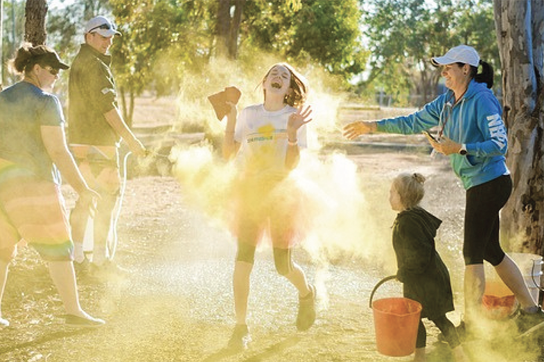 Eine Gruppe Menschen genießt ein Farbfestival im Freien, während gelbes Puder um ein lachendes Mädchen herum aufsteigt.
