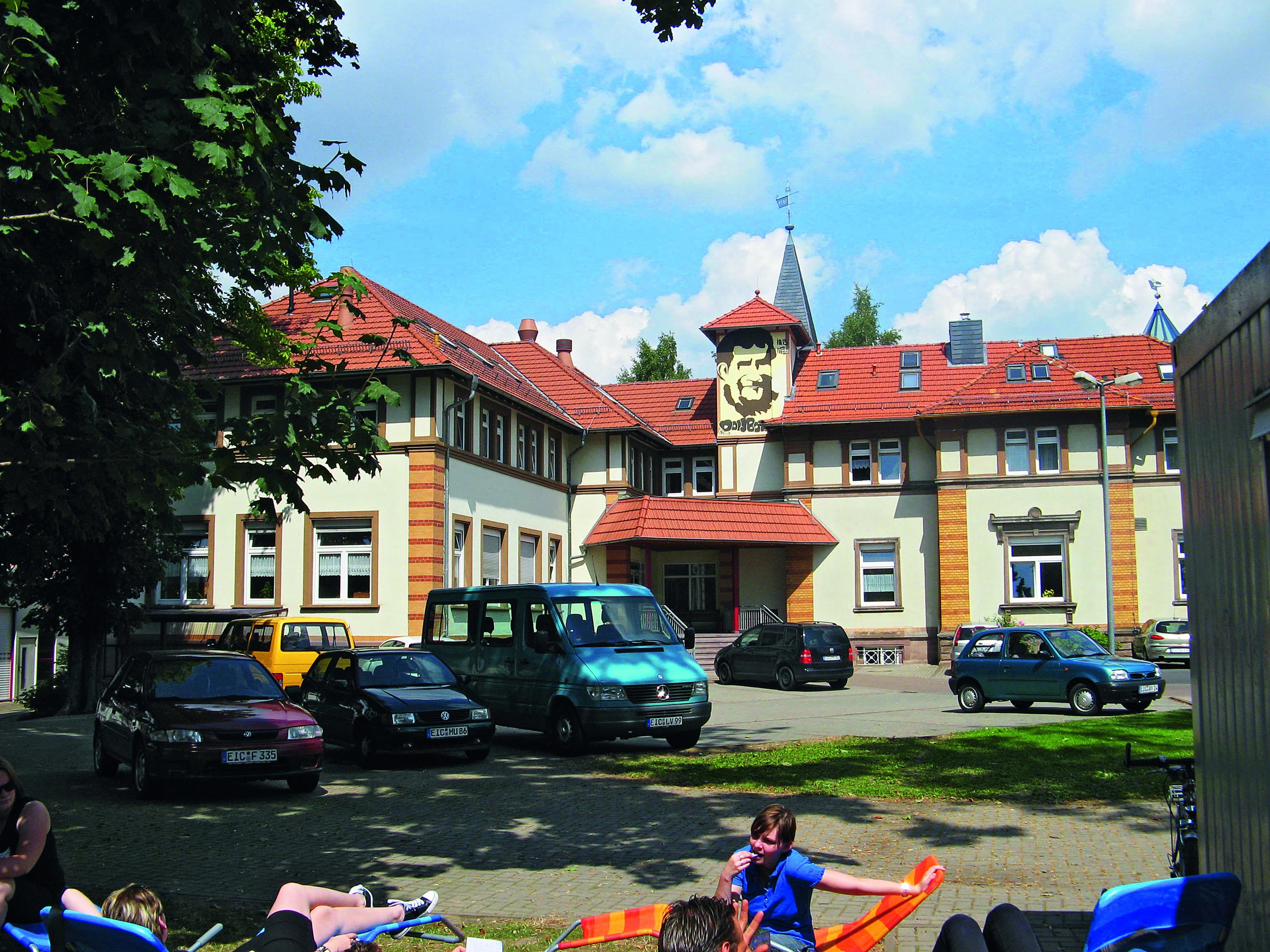 Gebäude von vorne mit Autos, Wiese und Kindern, Gebäude ist weiß / gelb das Dach rote Ziegelsteine