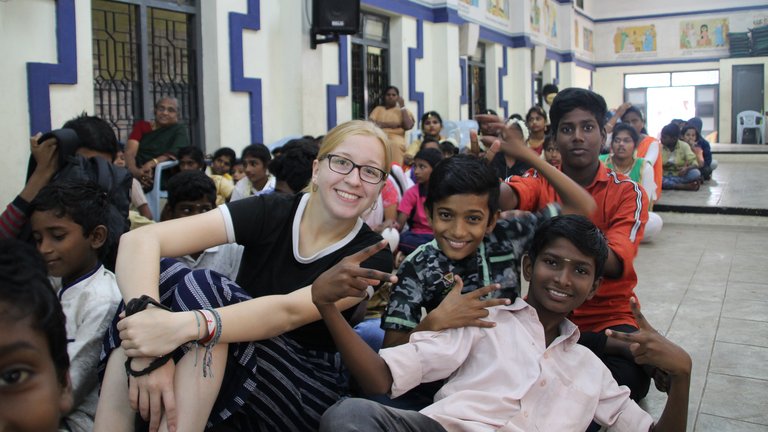 Gruppenfoto einer lächelnden blonden Frau und fröhlichen indischen Kindern, einige machen Friedenszeichen, in einer bunten Halle.