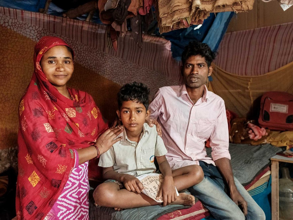 Porträt einer dreiköpfigen Familie: Frau in Rot, Mann in Rosa und ein Junge mit Papier, sitzend in ihrem Zuhause.
