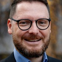 Nahaufnahme eines lächelnden Mannes mit dunklen Haaren, Bart und Brille, der ein kariertes Hemd und eine Jacke trägt.