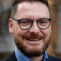 Nahaufnahme eines lächelnden Mannes mit dunklen Haaren, Bart und Brille, der ein kariertes Hemd und eine Jacke trägt.