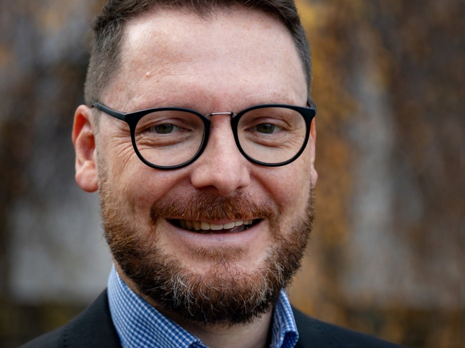 Nahaufnahme eines lächelnden Mannes mit dunklen Haaren, Bart und Brille, der ein kariertes Hemd und eine Jacke trägt.