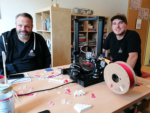 Zwei Männer präsentieren einen 3D-Drucker mit rotem Filament und kleinen gedruckten Objekten auf einem Tisch, inkl. Spardose.