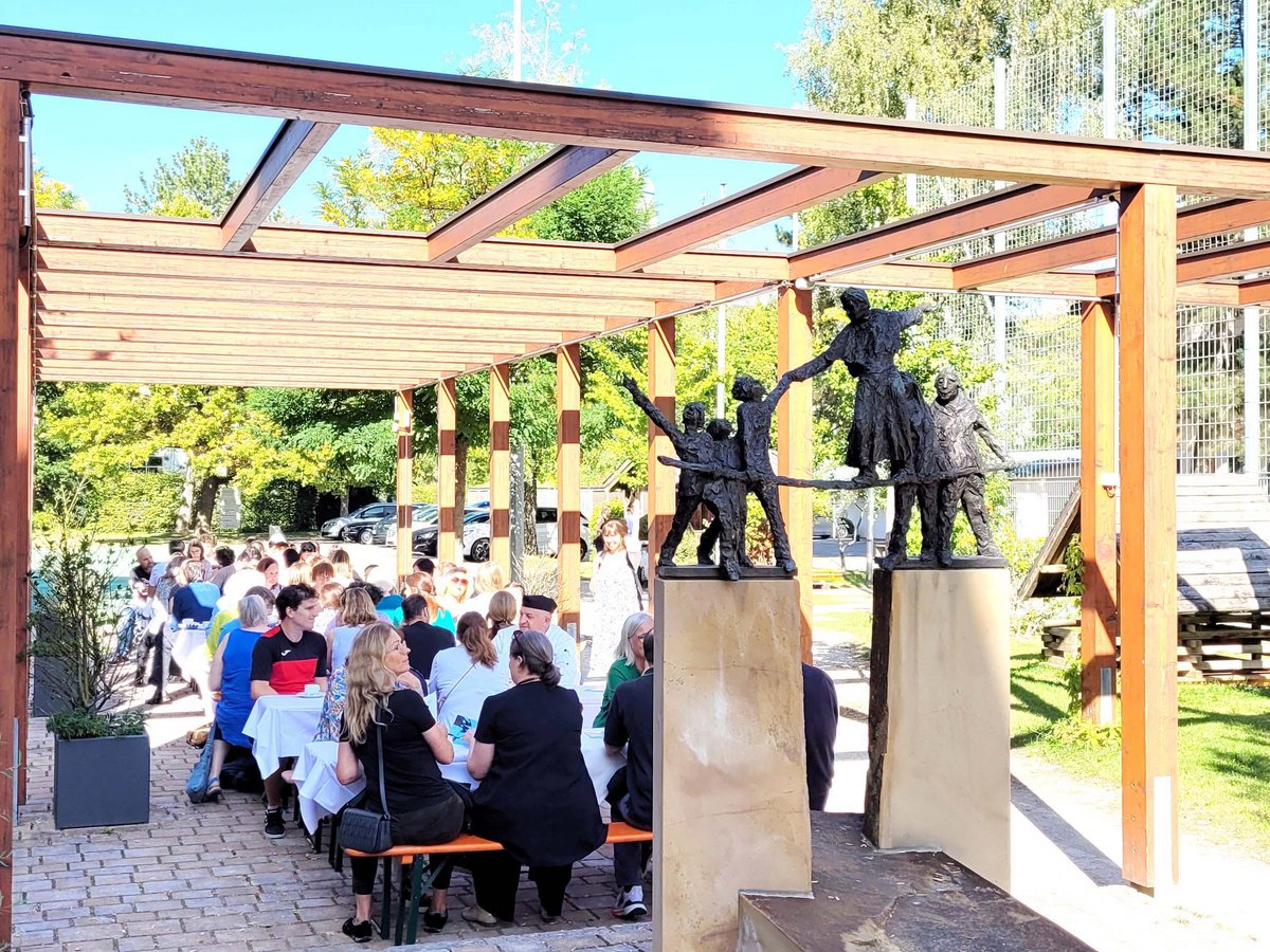 Menschen sitzen an Tischen unter einer Holzpergola mit einer Skulptur tanzender Figuren an einem sonnigen Tag.