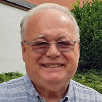 Ein freundlicher älterer Mann mit grauem Haar und Brille lächelt direkt in die Kamera, trägt ein blau-weiß gestreiftes Hemd.