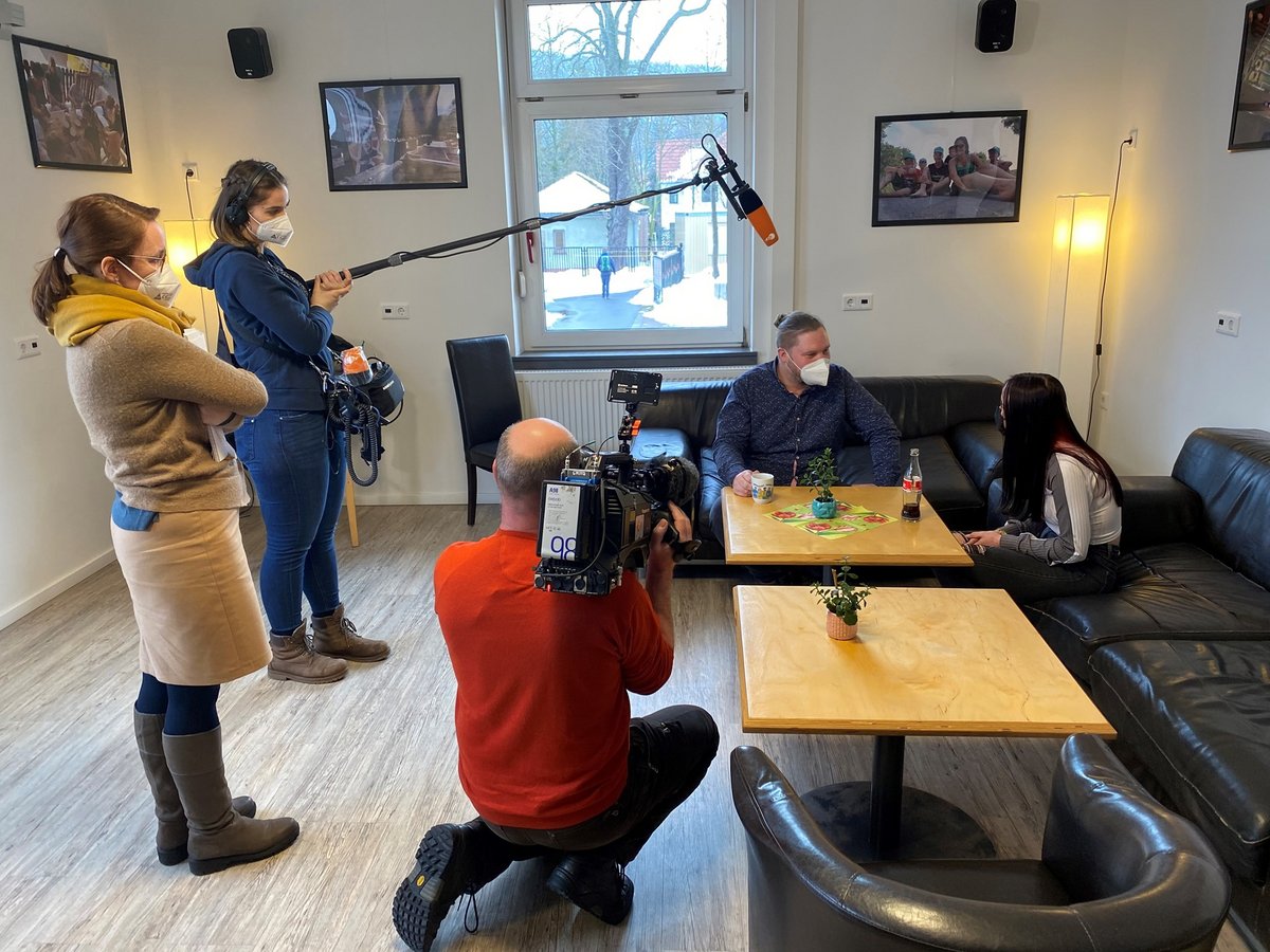 Ein Filmteam interviewt einen Mann und eine Frau mit Masken auf einem Sofa, mit Kameramann, Tontechniker und Regisseur.