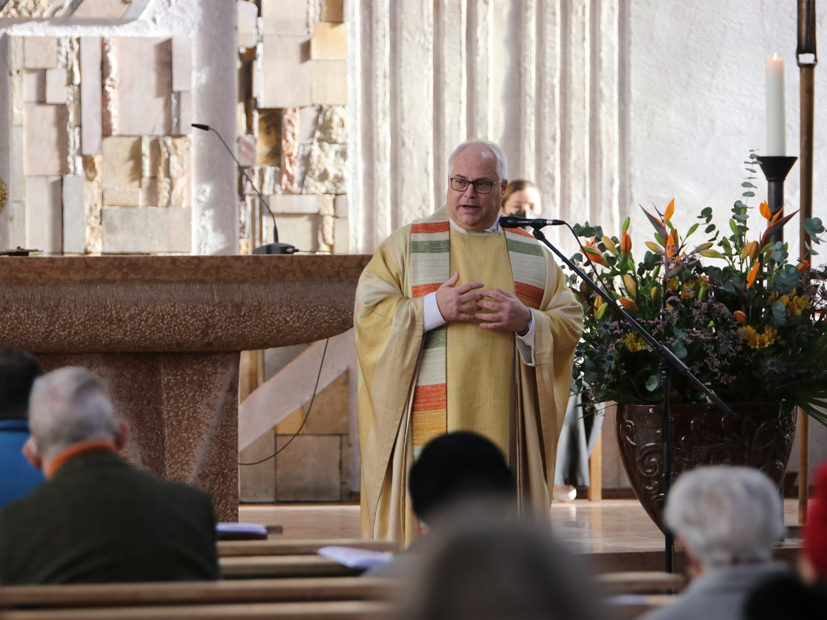 Ein Priester in goldenen Gewändern spricht an einem Altar in einer Kirche, mit Monstranz, Blumen und Gemeindemitgliedern.