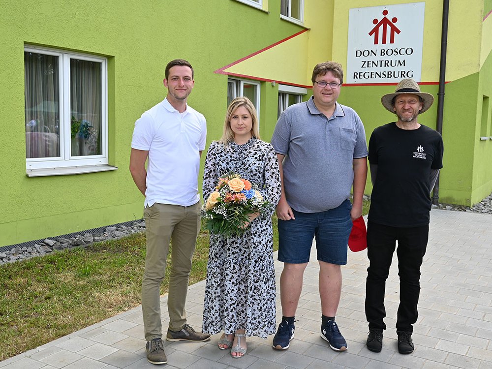 Vier Personen, darunter eine Frau mit Blumenstrauß, stehen vor dem lindgrünen Gebäude des Don Bosco Zentrums Regensburg.