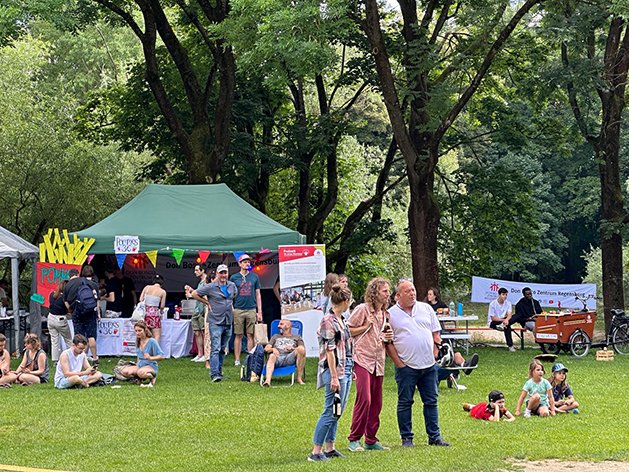 Ein Sommerfest oder Markt im Freien in einem Park mit vielen Menschen, Essensständen und Bäumen. Ein grünes Zelt ist zentral.