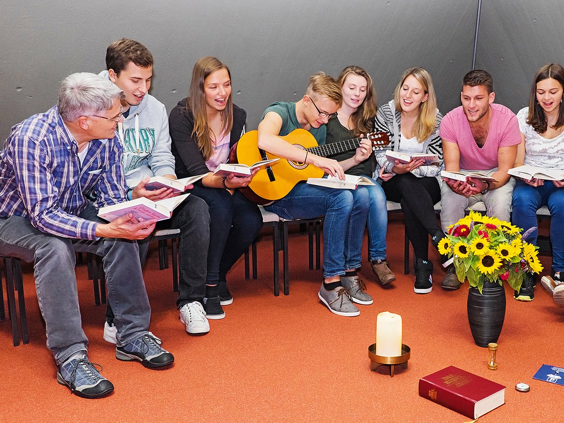 Neun Personen, darunter ein älterer Mann, lesen Bücher im Halbkreis. Ein junger Mann spielt Gitarre. Blumen, Kerze, Bibel im Vordergrund.
