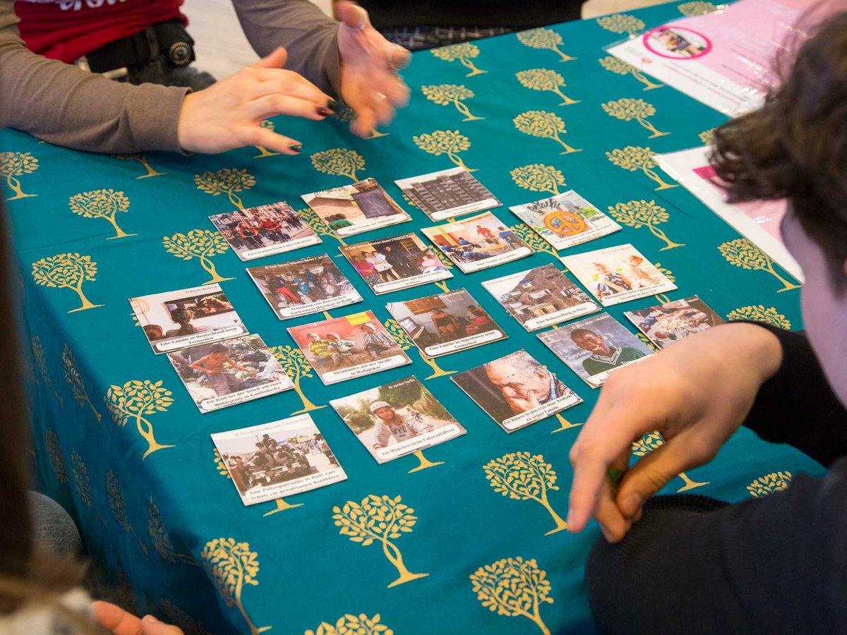 Hände spielen ein Kartenspiel auf einem türkisfarbenen Tisch mit gelben Bäumen. Karten zeigen globale Szenen mit deutschen Texten.