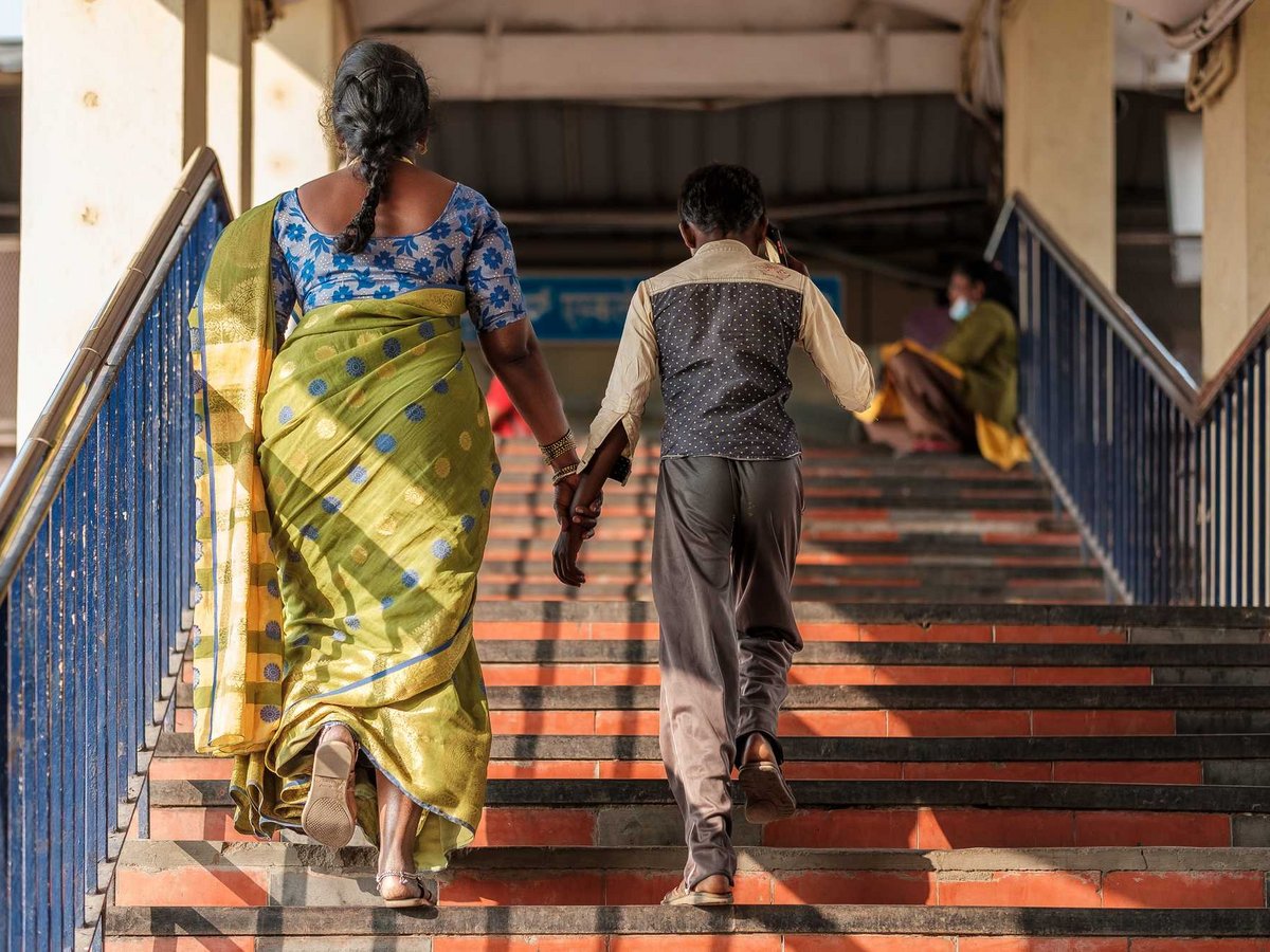 Rückenansicht einer Frau im grünen Sari und eines Jungen, die Hand in Hand eine Treppe mit blauem Geländer hinaufgehen.