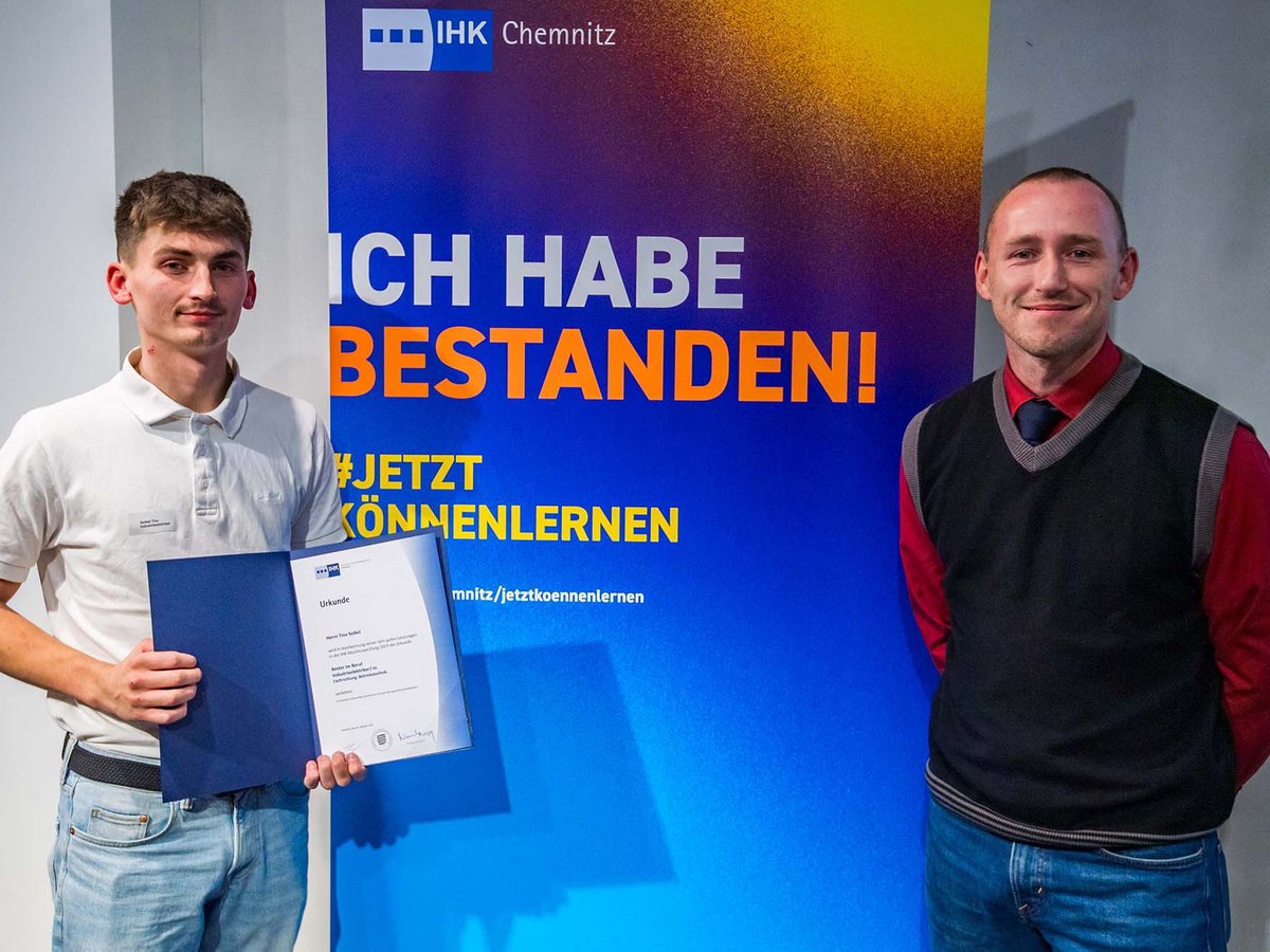 Zwei Männer mit Urkunde vor einem IHK Chemnitz Banner, das eine bestandene Prüfung feiert.