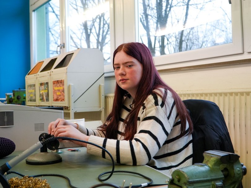 Junge Frau mit langen rötlichen Haaren in gestreiftem Pullover lötet an einer Werkbank und blickt in die Kamera.