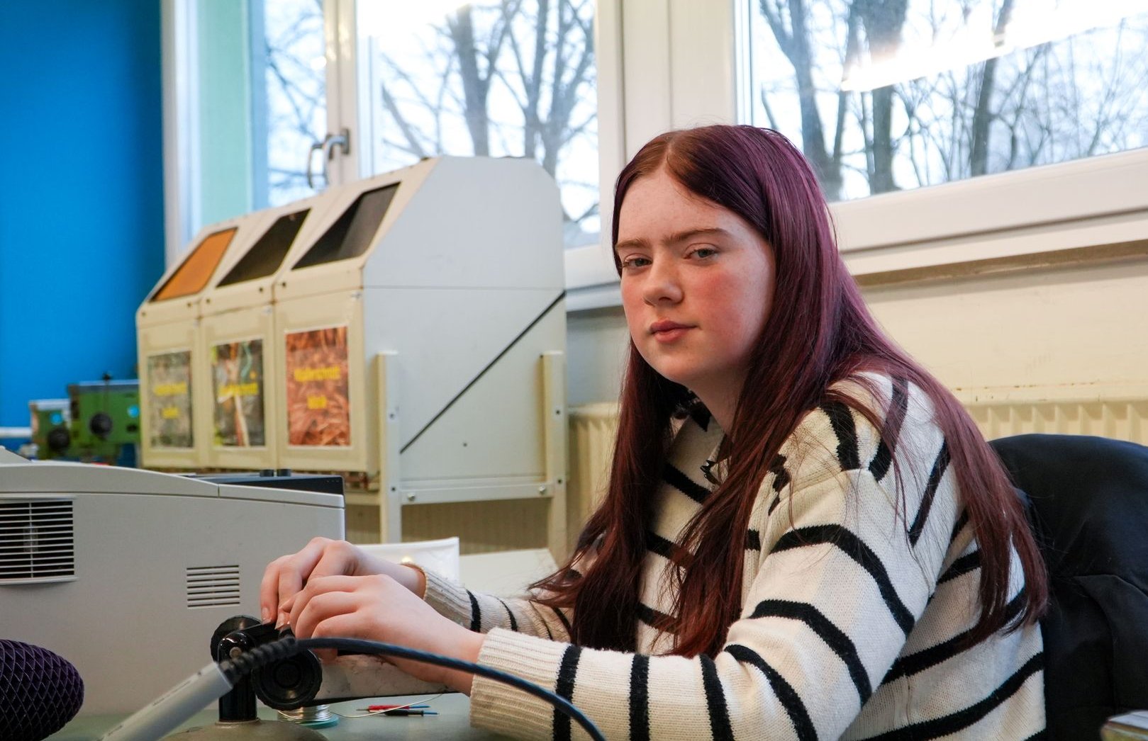 Junge Frau mit langen rötlichen Haaren in gestreiftem Pullover lötet an einer Werkbank und blickt in die Kamera.