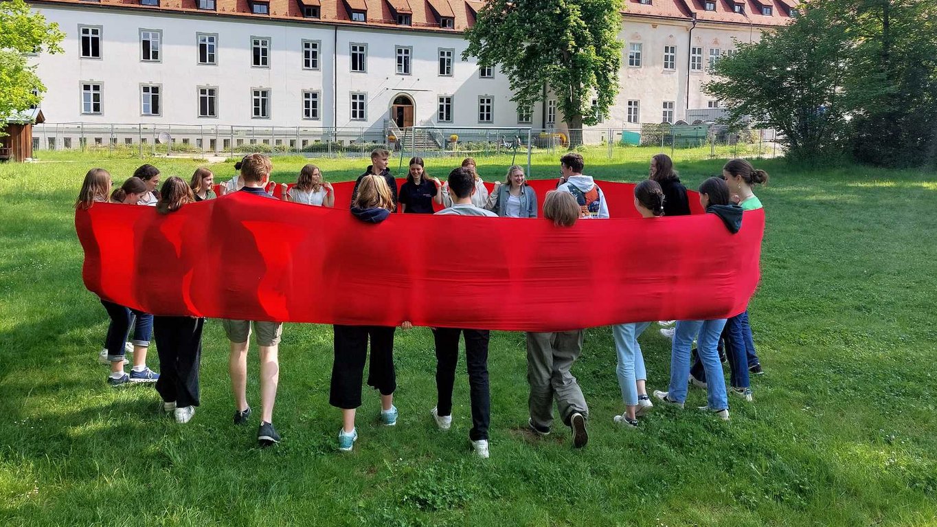 Eine Gruppe Jugendlicher bildet auf einer Wiese einen Kreis, indem sie ein langes rotes Tuch halten. Im Hintergrund ein großes Gebäude.