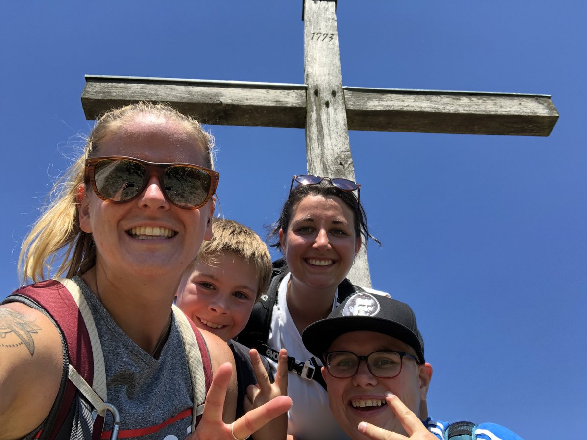 Vier Personen lachen in die Kamera, hinter ihnen ein Gipfelkreuz