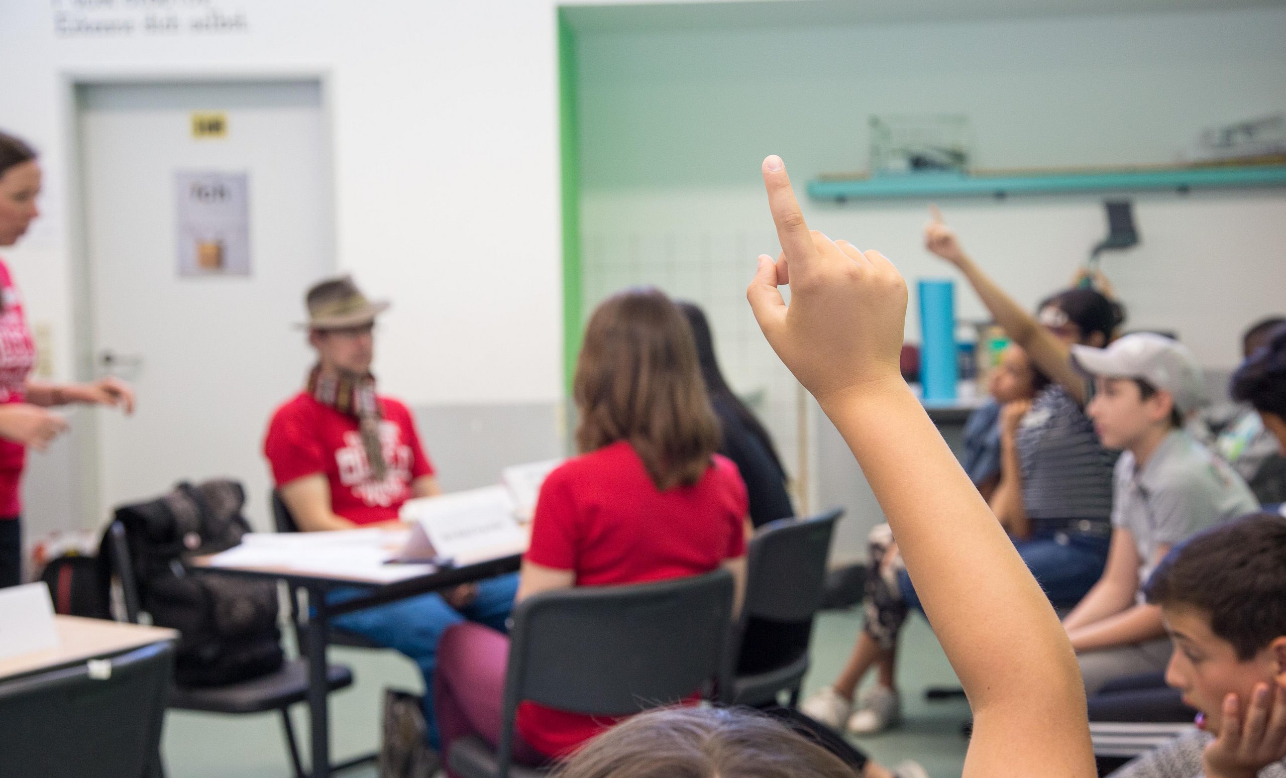 Erhobene Hand eines Kindes im Klassenzimmer mit anderen teilnehmenden Schülern und einem Erwachsenen im Hintergrund.