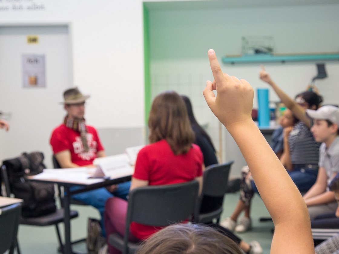 Erhobene Hand eines Kindes im Klassenzimmer mit anderen teilnehmenden Schülern und einem Erwachsenen im Hintergrund.