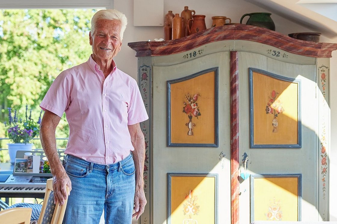 Lächelnder älterer Mann in rosafarbenem Hemd und Jeans, steht neben einem antiken, bemalten Schrank in einem hellen Raum.