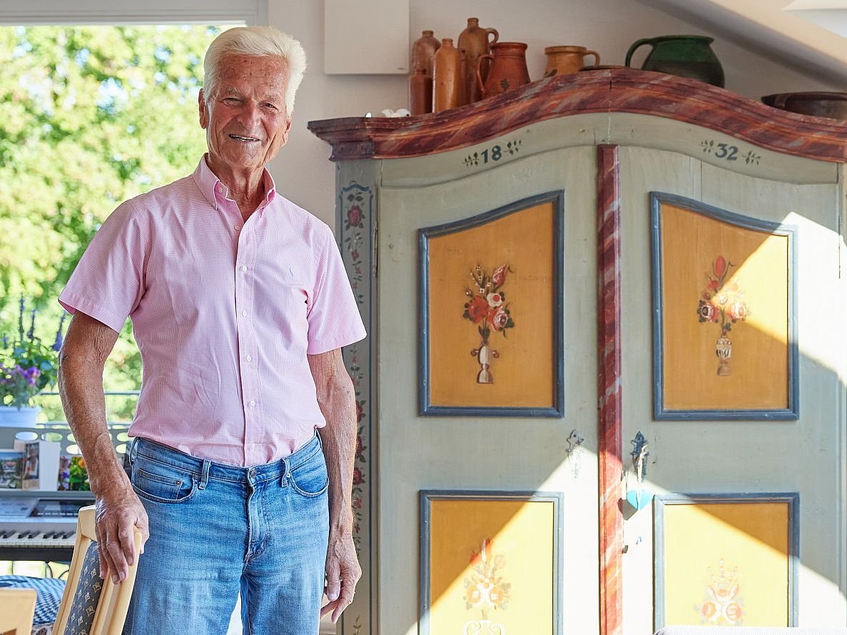 Lächelnder älterer Mann in rosafarbenem Hemd und Jeans, steht neben einem antiken, bemalten Schrank in einem hellen Raum.