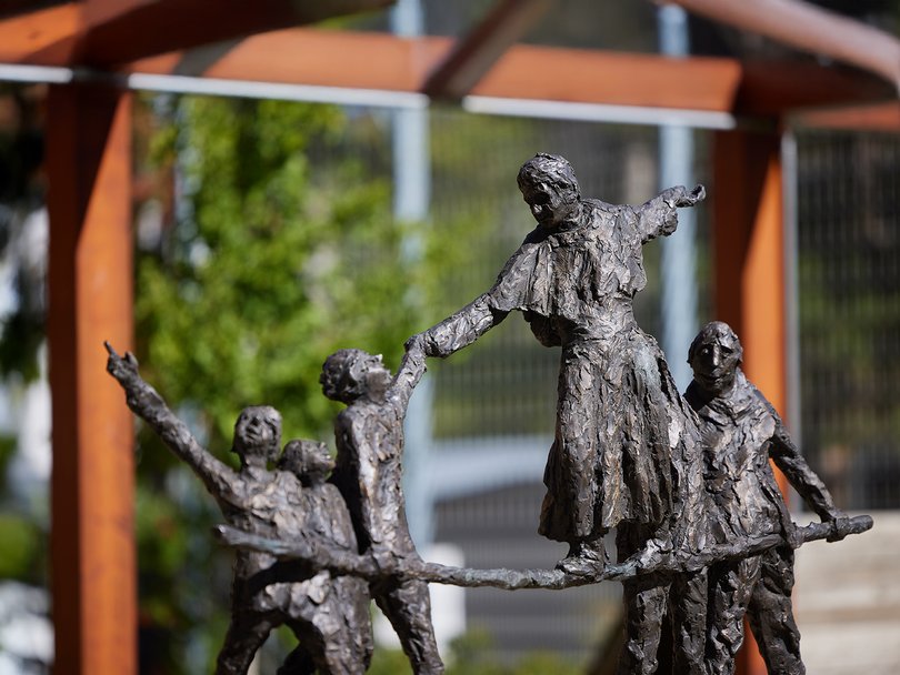 Texturierte Bronzeskulptur mehrerer Figuren, einige zeigen und reichen, vor verschwommenem Außenhintergrund.