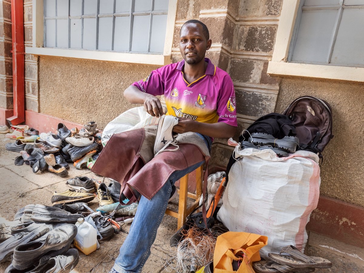 Ein lächelnder Schuhmacher in lila Hemd und Lederschürze repariert im Freien einen weißen Schuh, umgeben von Schuhstapeln und Werkzeugen.