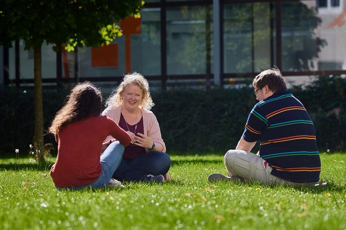 Drei Personen sitzen an einem sonnigen Tag draußen auf einer Wiese, eine Frau lächelt eine andere während eines Gesprächs an.