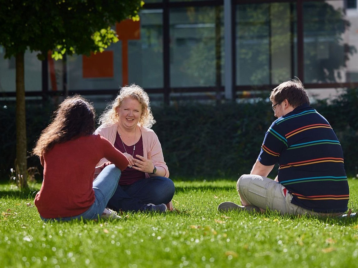Drei Personen sitzen an einem sonnigen Tag draußen auf einer Wiese, eine Frau lächelt eine andere während eines Gesprächs an.