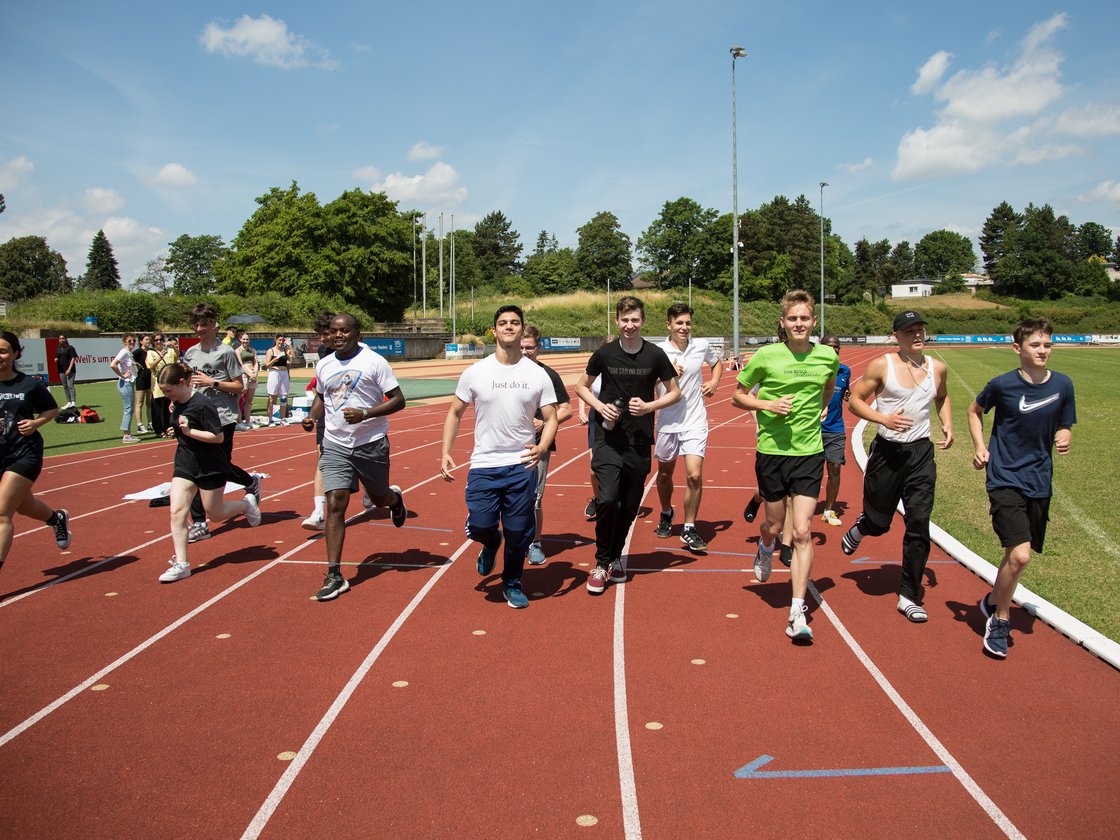 Vielfältige Gruppe junger Menschen läuft an einem sonnigen Tag auf einer roten Leichtathletikbahn.