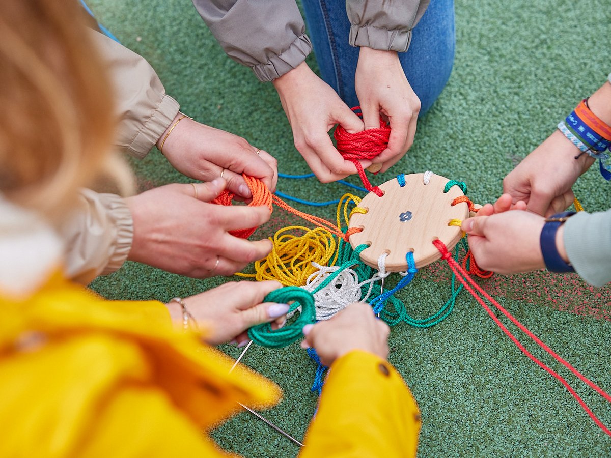 Hände mehrerer Personen flechten gemeinsam bunte Seile durch eine Holzscheibe auf grünem Untergrund.