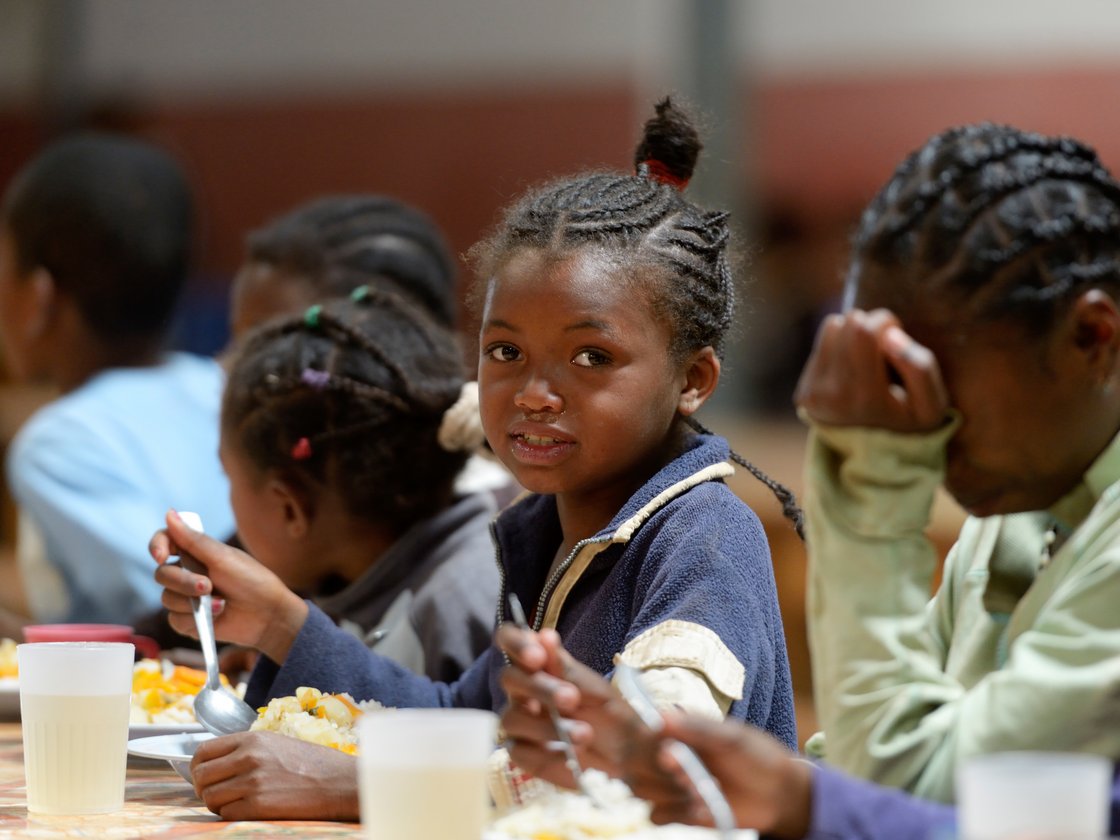 Ein junges afrikanisches Mädchen mit geflochtenen Haaren blickt in die Kamera, während es mit anderen Kindern isst.