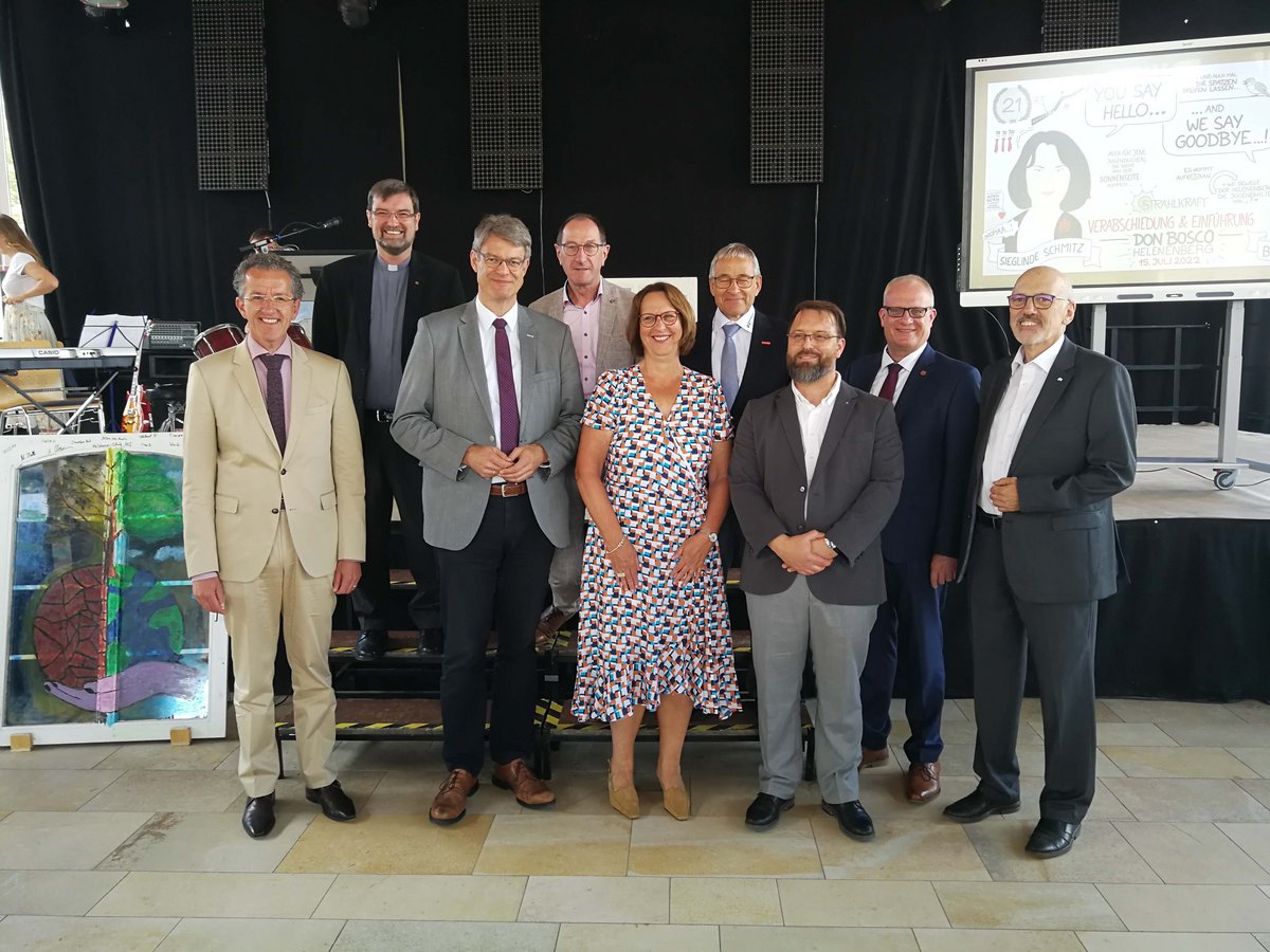 Neun festlich gekleidete Personen stehen bei einer Verabschiedungs- und Einführungsveranstaltung auf einer Bühne.