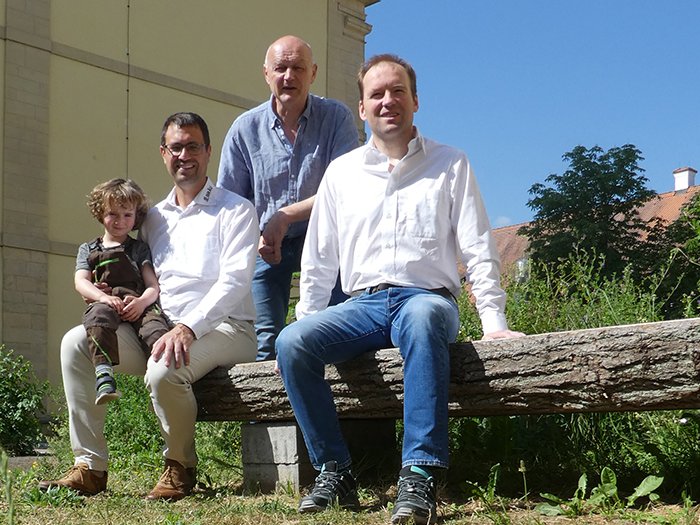 Drei Männer und ein Kind sitzen an einem sonnigen Tag draußen auf einer Holzbank, mit einem Gebäude und Grün im Hintergrund.