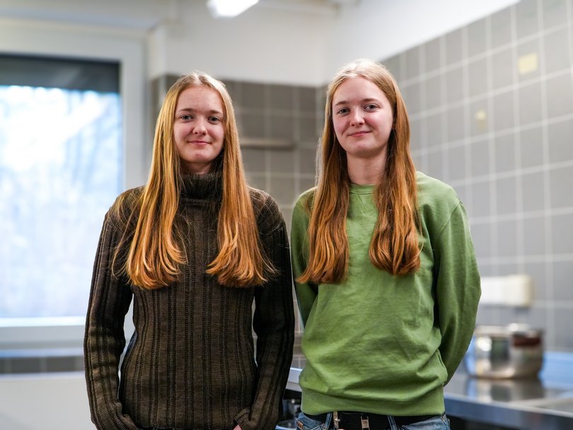 Zwei lächelnde junge Frauen mit langen blonden Haaren, möglicherweise Zwillinge, stehen nebeneinander in einem Innenraum.