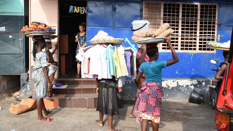 Marktszene mit Menschen, die Waren wie Tomaten, Yamswurzeln und Kleidung auf dem Kopf tragen.