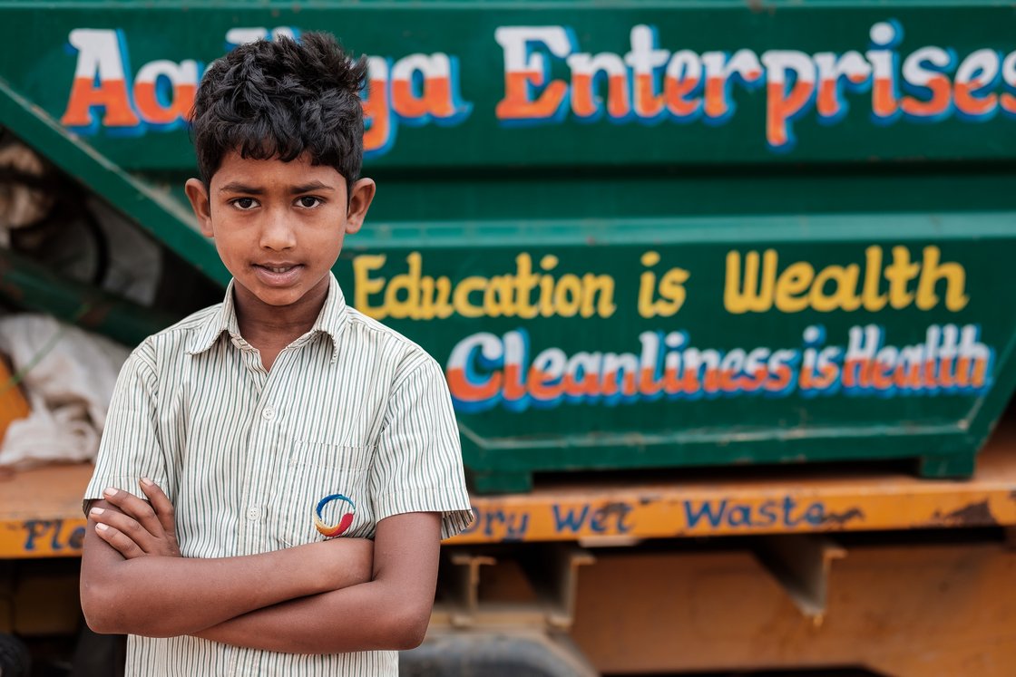 Junger indischer Junge mit verschränkten Armen vor einem grünen Hintergrund mit der Aufschrift 'Education is Wealth'.