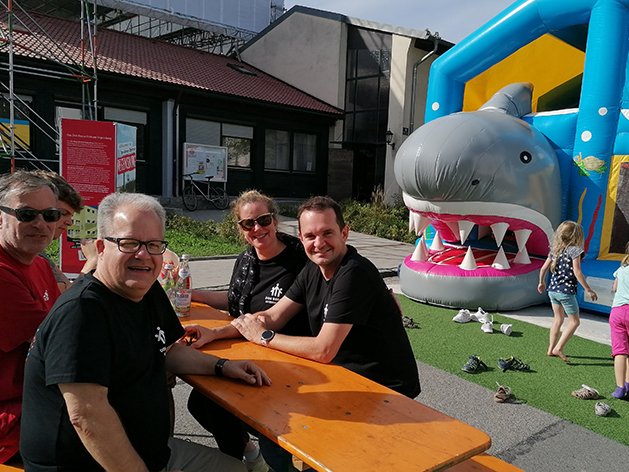 Mehrere Erwachsene sitzen lächelnd an einem Holztisch, während Kinder neben einer aufblasbaren Haifisch-Hüpfburg spielen.