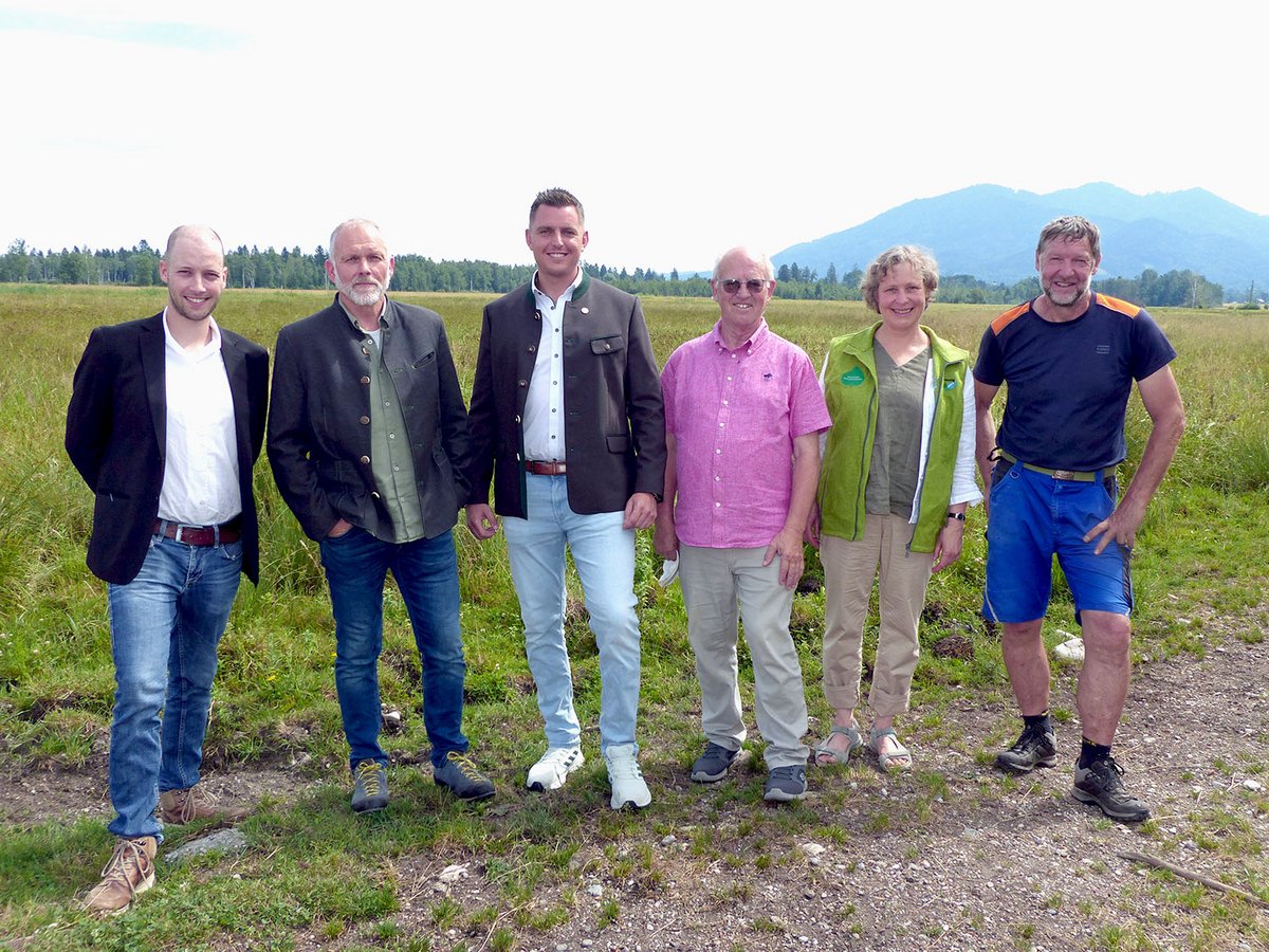 Sechs Personen, fünf Männer und eine Frau, in traditioneller und legerer Kleidung, stehen auf einer Wiese vor Bergen.