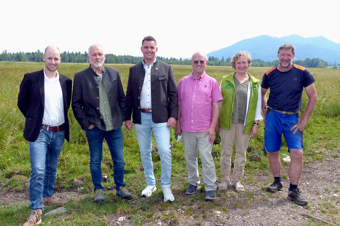 Sechs Personen, fünf Männer und eine Frau, in traditioneller und legerer Kleidung, stehen auf einer Wiese vor Bergen.