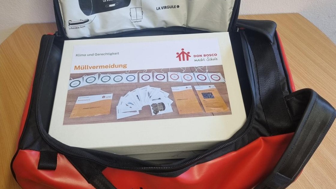Rote und schwarze HORS-BORD BAG mit offenem Deckel, in der ein Karton zum Thema "Müllvermeidung" von Don Bosco liegt.