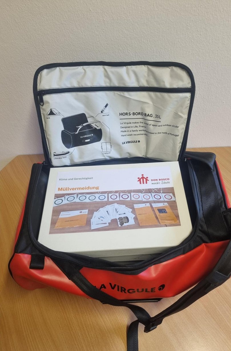 Rote und schwarze HORS-BORD BAG mit offenem Deckel, in der ein Karton zum Thema "Müllvermeidung" von Don Bosco liegt.