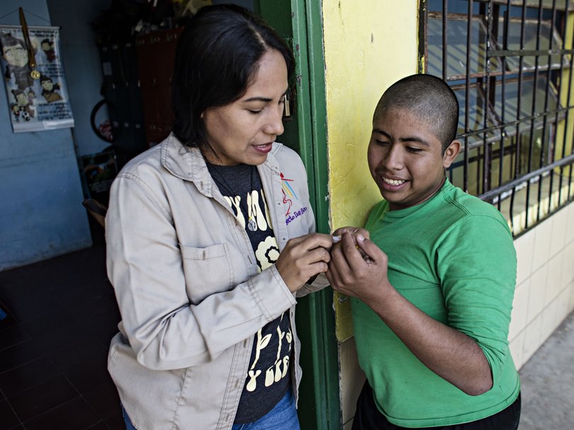 Eine Frau und ein Junge tauschen lächelnd etwas Kleines in einer Türöffnung aus, im Hintergrund eine gelbe Wand mit Gitterfenster.